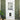 A white front door with a small pet door and glass panes, flanked by two green trellis panels with climbing plants. A woven rug lies on the concrete floor, with potted plants at the lower left.