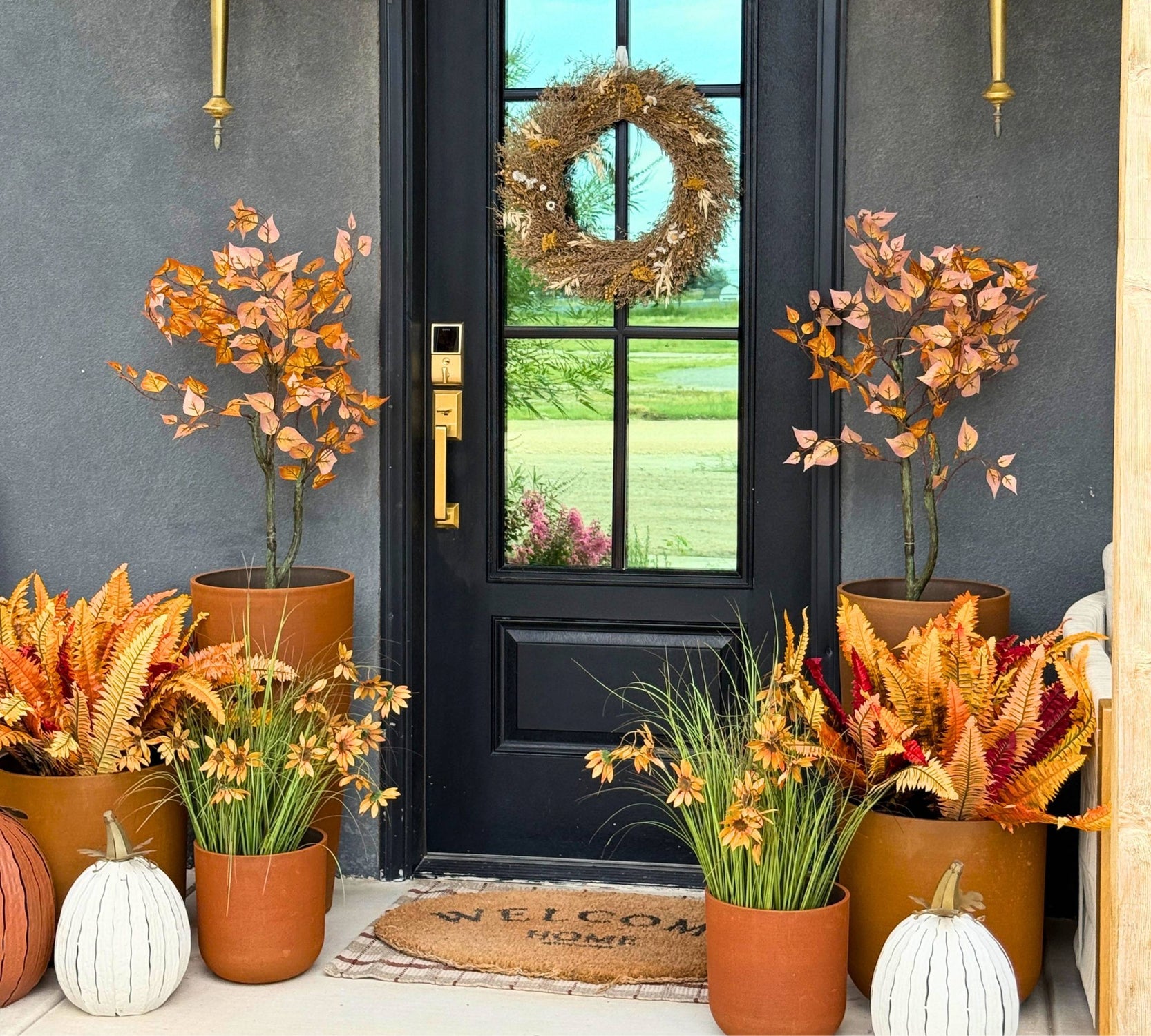 An autumn-themed front porch display with multiple terracotta pots containing orange, yellow, and burgundy faux foliage, ferns, and decorative pumpkins flanking a black door with a woven wreath.